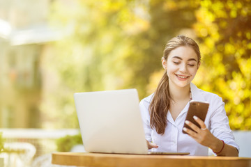 Young smiling businesswoman sitting at table in coffee shop outdoors and uses smartphone on table cup of coffee and notebook
