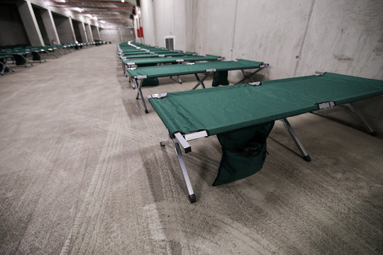 Camp folding cots are being set up in the underground parking of a stadium