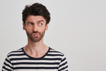 Clouse up of young brunet with beard looking to the right with distrust and discontent, frowning brows, lips pursed wears black and white striped tshirt, stands leftside isolated over white background