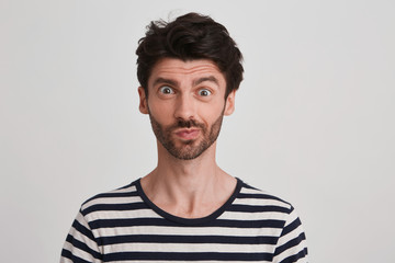 Clouse up of young brunet with beard looking suddenly surprised and discontent, eyebrows raised, lips pursed wears black and white striped tshirt, stands rightside isolated over white background
