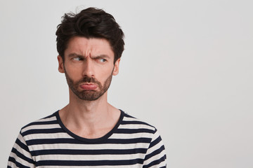 Clouse up of young brown hair man with beard looks sad and offended, head turned right frowned eyebrows, lips pout, wears black and white striped tshirt, stands leftside isolated over white background