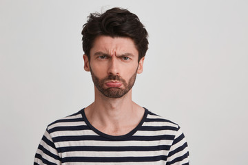 Obraz premium Clouse up of young brown hair man with beard looks sad and offended, frowned eyebrows, lips pout, wears black and white striped tshirt, isolated over white background
