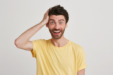 Obraz premium Portrait of happy and very excited attractive young brunet with beard eyebrows raised, keeps one hand on head, wears yellow casual tshirt, isolated over white background