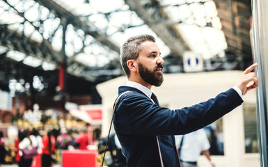 Hipster businessman standing on bus or train station, checking timetable.