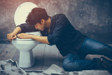 Asian handsome man showing symptoms drunk heavily in the toilet. He sat beside the toilet bowl, a stance vomiting and losing consciousness after drinking alcohol, beer, vodka with a friend in a pub. - obrazy, fototapety, plakaty