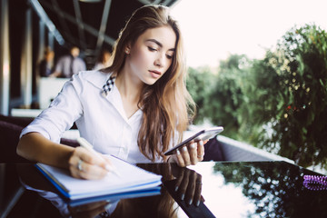 Young woman student in eyeglasses concentrated on writing plan in notepad while sending messages on mobile. Pensive hipster girl in eyewear noting information from website browsed on smartphone