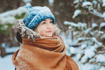 winter portrait of happy young woman walking in snowy forest in warm outfit, knitted hat and oversize scarf. Exploring nature on winter and Christmas vacations concept