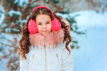 Winter portrait of happy kid girl in pink earmuffs walking outdoor in snowy winter forest. Happy childhood and active winter holidays concept
