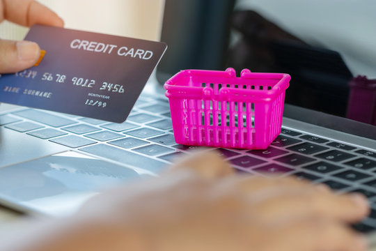 Empty pink basket and hand holding credit card mockup on laptop keyboard. Consumer can buy product anywhere anytime from seller using web browser or application. Online shopping and e-commerce concept