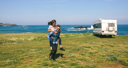 Young man piggybacking his girlfriend near the coast with camper van in the background