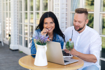 A happy married couple is resting in a cafe, laughing, enjoying delicious cocktails, using smartphones and a laptop. The girl drinks her drink and straws, the man is distracted by the SMS from work