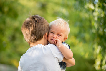 Little toddler baby boy, crying, older brother carrying him in the park, baby looking at camera