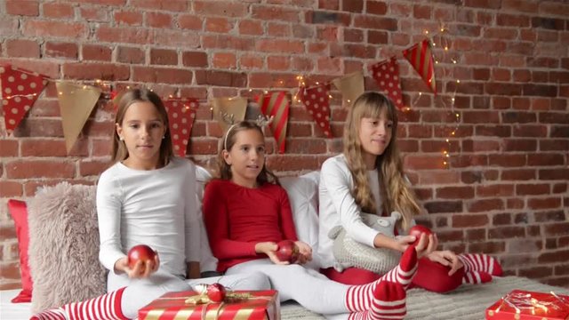 Three sisters sitting on the bed in soft pajamas and playing with red apples. The bedroom is decorated with Christmas lights. Merry Christmas and a Happy New Year.