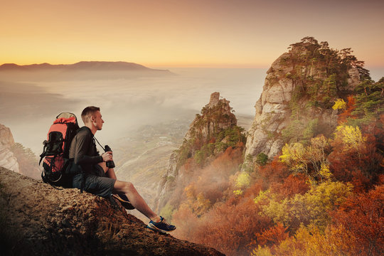 Young man hiker with backpack and binoculars sitting on a cliff and enjoying a Mountain view.