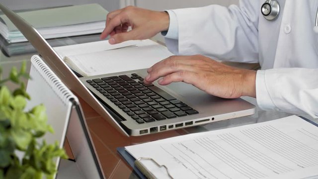 Close-up view of unrecognizable male doctor typing on laptop and taking notes in medical record of patient while sitting at table in consulting room