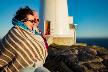Loving mature couple hiking, sitting on windy top of rock with lighthouse. Mature man and woman wrapped in blanket, hugging and Happily smiling. Scenic view of sea, mountains. Norway, Lindesnes.