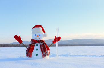 Obraz premium The snowman in plaid scarf, red hat, gloves and icicle in the hand. Nice landscape with the mountains on the background. Field covered with snow. Cold winter day.
