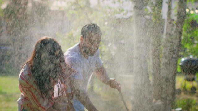 Happy Young Couple Has Fun On A Hot Summer Day Playing With Water Hose Sprinkler In The Garden. Two Young People In Love Got Wet Jokingly Fighting With Hose. In Slow Motion.