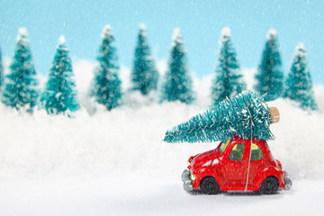Red toy car carrying a Christmas tree through the forest in snowfall. Cristmas and new year background.