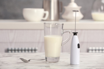 Milk frother device, glass cup and spoon on table in kitchen