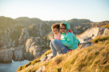 Naklejka na ściany i meble Two little girls play on rocky northern seashore. Sit, laugh, hug, explore the coastal rocks. Travel and enjoy a great adventure in Norway. Beautiful view of fjord and mountains in sunset.