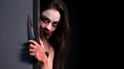 Makeup for halloween party. A girl with a bloodied knife in her hand looks out from behind a gray...