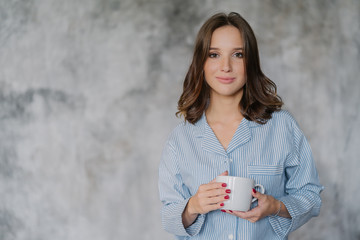 Attractive woman dressed in nightwear, holds white mug with coffee or tea, has morning drink, poses indoor against blurred background with copy space for your advertisement or promotional content © VK Studio