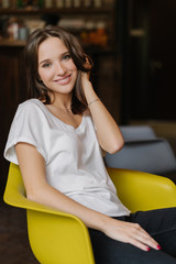 Obraz premium Vertical shot of pretty girl dressed in white t shirt and jeans, has happy look at camera, sits at yellow armchair, being in good mood, feels carefree, has pleasant smile on face. Feminity and rest
