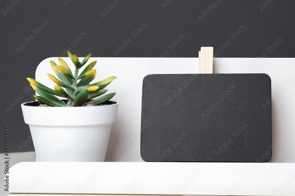 Poster black clip blackboard on modern pencil box and green plant in white pot on table and dark grey wall 