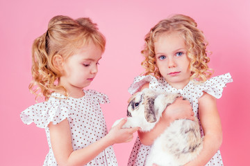 two cute little blonde girl with a bunny rabbit easter in studio pink background.beautiful female kids celebrating Easter Holiday with pets,child painted eggs in wicker basket.dream birthday present