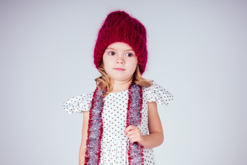 Beautiful little girl in dress and red knitted hat in studio on white background. Happy little smiling female child with shiny Christmas tinsel. Christmas miracles Happy New Year