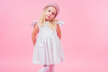body portrait of adorable little girl blonde girl blue eyes model in beige knitted pink hat in white princess dress in peas autumnal springtime season in studio shot on pink background