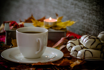 white cup and saucer with tea or coffee is on the table, maple leaves, next to it is a plate with white cookies with chocolate stripes, a book with a lit small candle in the background, dark gray tone