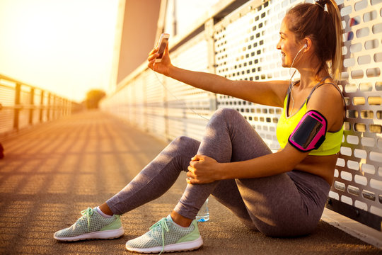 Female Runner Resting After Running On Bridge, Taking Selfie With Mobile Phone