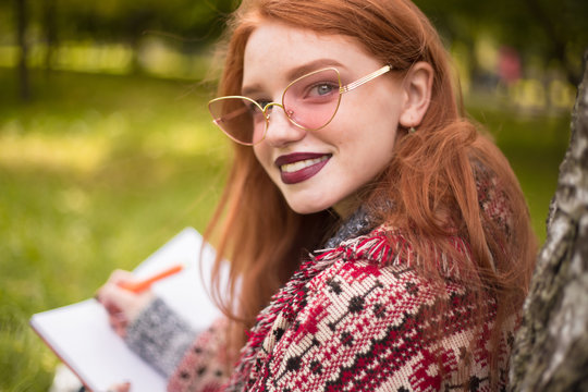 Woman with freckles and dark lipstick wearing sunglasses reading book writing notes doing homework outdoors in park sit under big tree on grass looking camera.