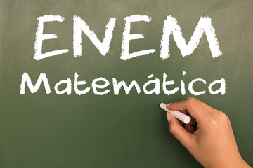 Female hand writing with chalk on empty green chalkboard. (translation portuguese: ENEM - National High School Exam Brazil - mathematics)
