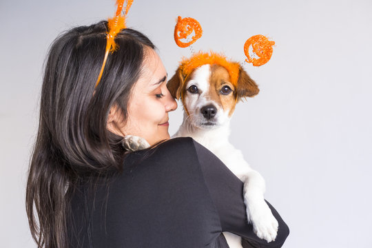 young woman holding her cute small dog over white background. Matching pumpkin diadems. Halloween concept. Indoors