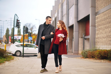 Young romantic couple, beautiful girl with flowers has dating in the city. Man in gray overcoat and model in red coat. Loving happy, smile, enjoy a walk together on a authmn day near shopping center. 