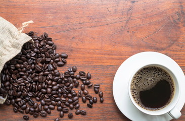 Top view of coffee beans in sack and black coffee in white cup on wooden table with copy space for text or image.
