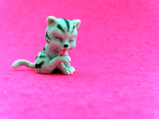 Closeup of plastic toy kitten licking his leg on a pink background