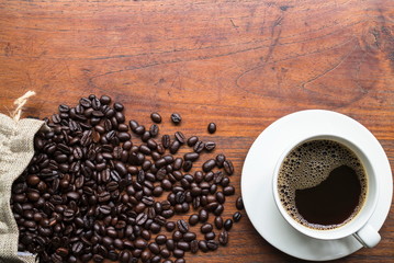 Top view of coffee beans in sack and black coffee in white cup on wooden table with copy space for text or image.