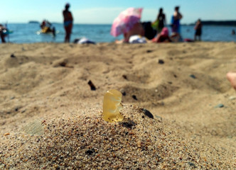 Closeup of a lonely yellow gummy bear on the sandy beach with blurred people in the background (selective focus)