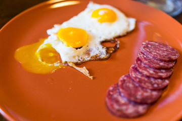 Closeup of two broken runny fried eggs on large orange plate with cured meat hard salami slices, yellow liquid