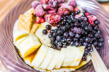 Macro closeup of oatmeal oat grain porridge bowl with sliced bananas, peanut butter, frozen berries, raspberries, blueberries fruit, maple syrup