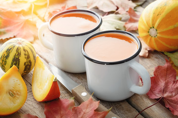 Two white enameled cups of pumpkin juice, pumpkins and fallen maple leaves on table.