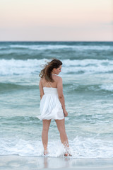 Fototapeta na wymiar Young woman back standing in white dress on beach sunset in Florida panhandle with wind, ocean waves crashing legs, tan skin, looking down vertical view
