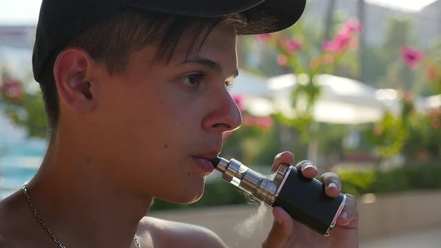  Profile of a young brunet man in a baseball cap toking an electronic cigarette trying to relax in a green park with sunshades in slow motion