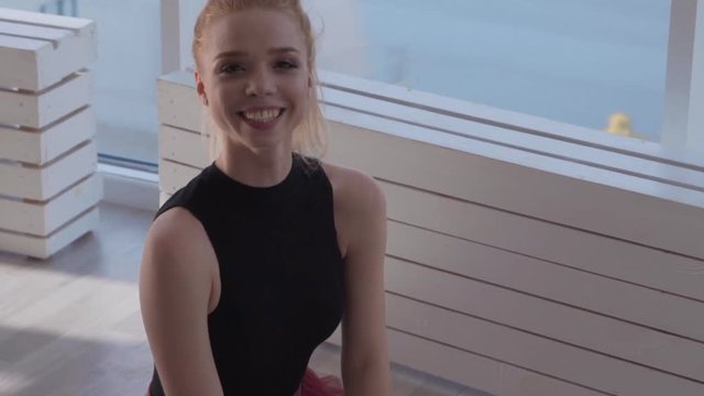 young tender girl ballerina sitting on the floor and laughing into the camera. pretty blonde with smooth hair in a black dance tights and a red skirt. slow motion
