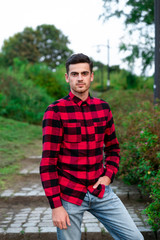 Naklejka na ściany i meble Portrait of stylish male student with bread posing outdoors, looking at camera. Handsome man wearing in red checked shirt walking in autumn park. Concept of study, and mans fashion.