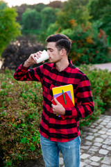 Fototapeta premium Happy student drinking hot coffee and resting after studying. Handsome and stylish brunet male wearing in red checked shirt walking in autumn park with notes and showing at camera cup of coffee. 